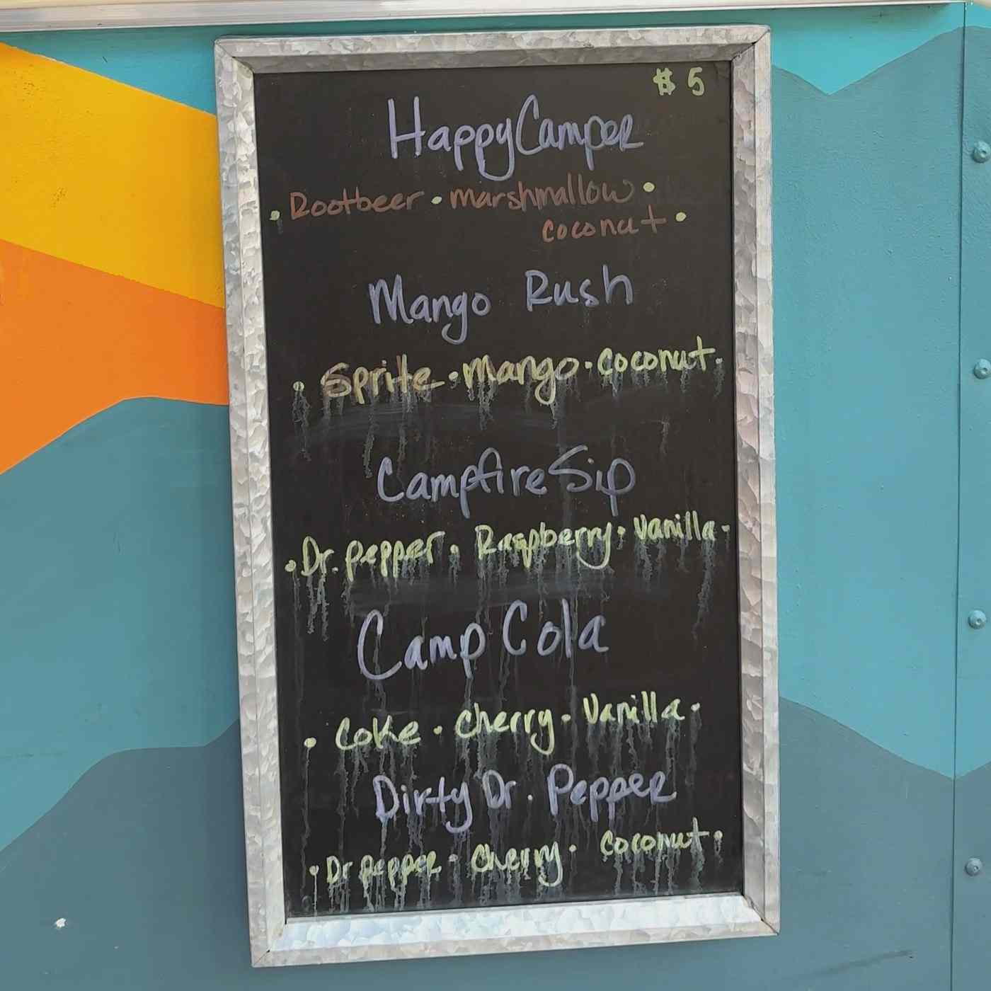 The camp soda chalkboard on the trailer: Happy Camper, Mango Rush, Campfire Sip, Camp Cola and Dirty Dr. Pepper