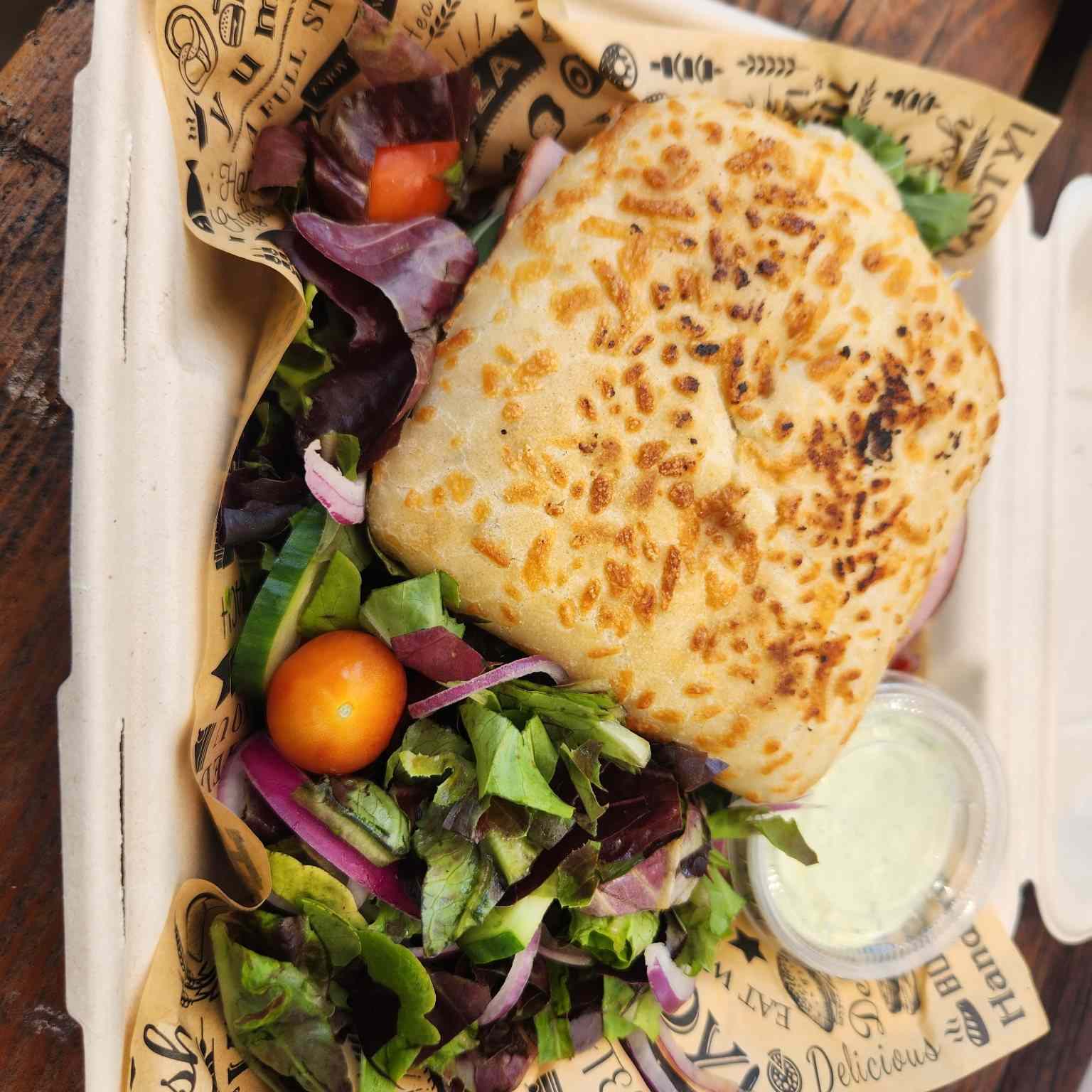 A ciabatta sandwich with mixed greens, cherry tomato and ranch in a takeaway box
